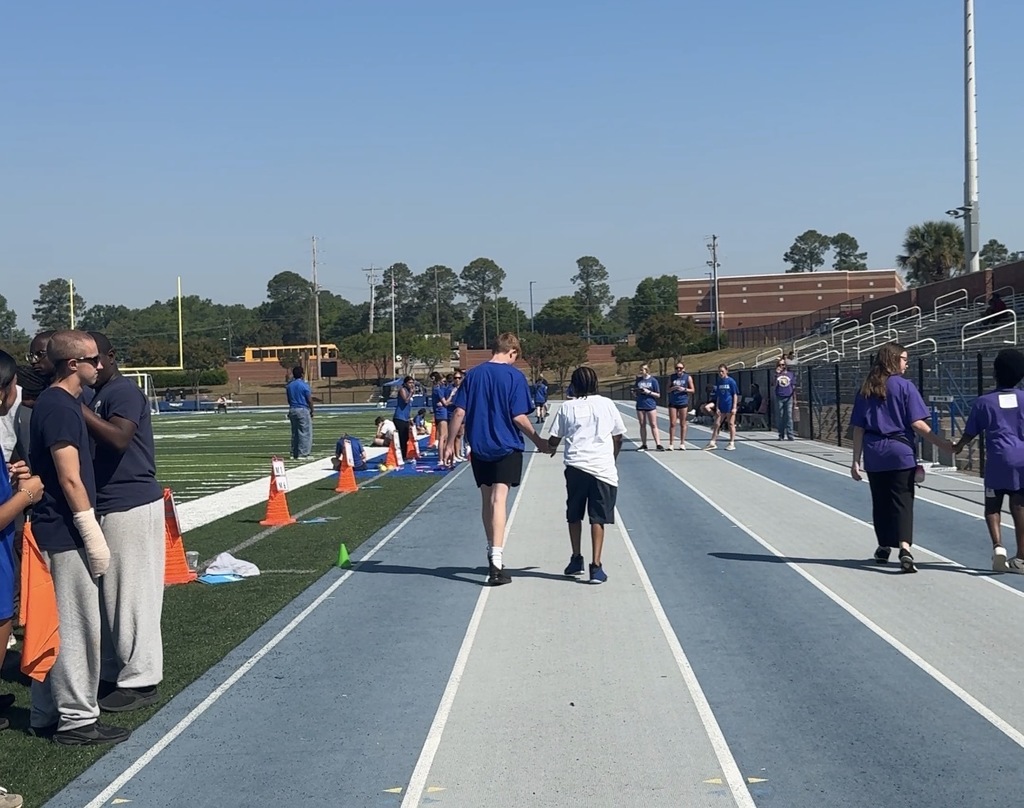Two students are holding hands and walking on the track. There backs are towards the camera. Other students and adults are seen on the sideline and track standing and walking.