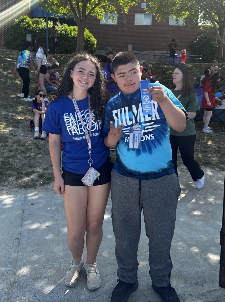 Two students stand side by side and look at the camera. One male student is holding his blue ribbon he received from placing in an event at the Olympics.