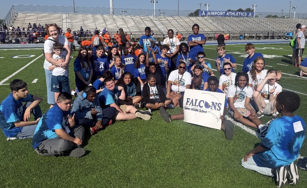 A group of students and adults sit and stand on a high school football field. They are all looking towards the camera. One student seated on the ground holds a Falcons Fulmer Middle School poster.