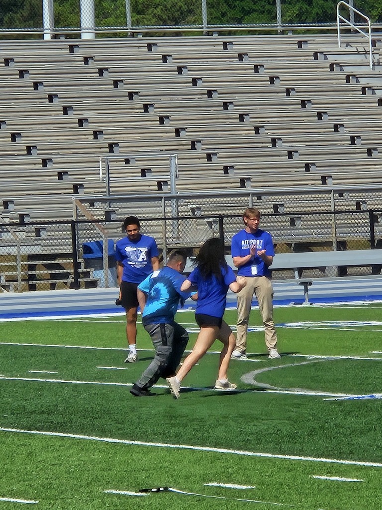 Two students run side by side on the football field. An adult and high school student are shown in the background watching them run and cheering them on by clapping.