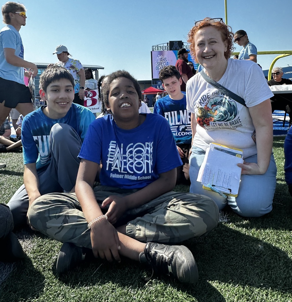 Three students and a teacher sit on the ground outside on the football field and smile at the camera.