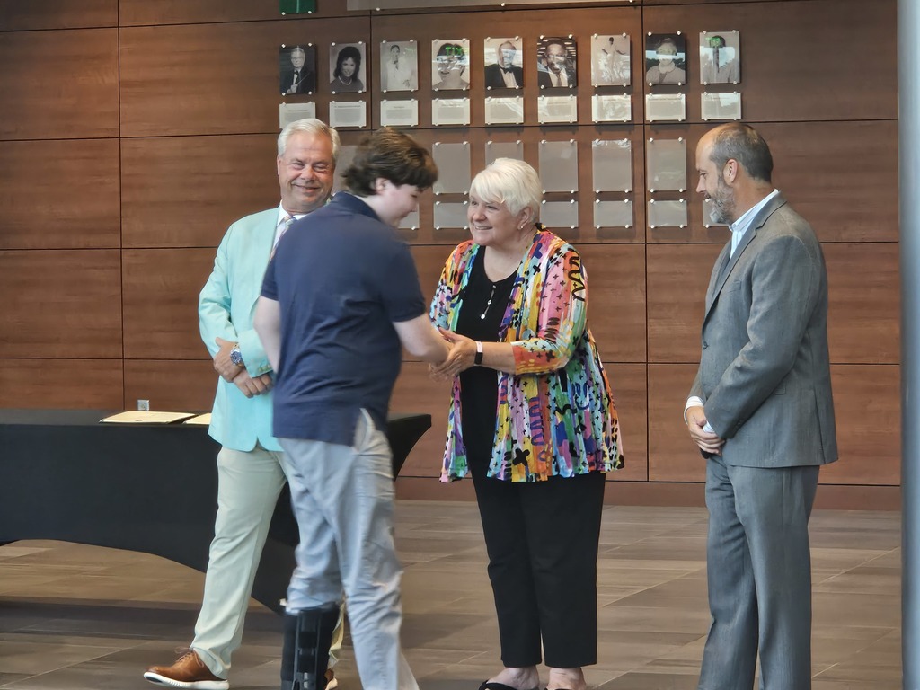 A student shakes hands with a board member. Other board members are shown standing side by side.