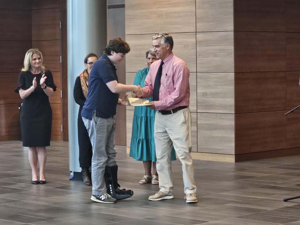A student shakes hands with the school principal and receives a certificate for being a Junior Scholar. Adults are shown standing in the background.