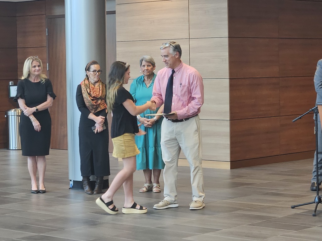 A student shakes hands with the school principal and receives a certificate for being a Junior Scholar. Adults are shown standing in the background.