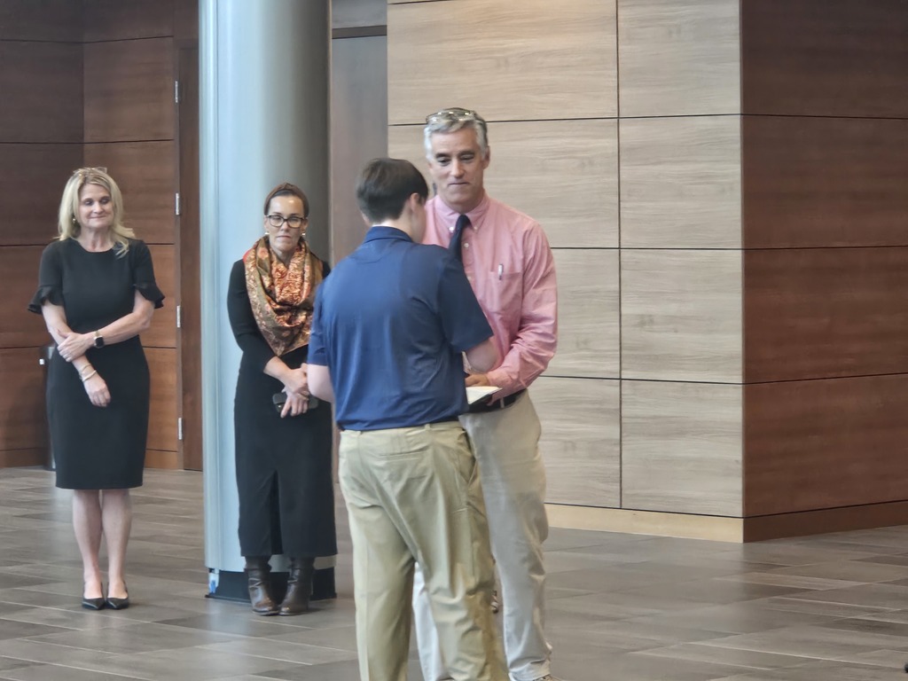 A student shakes hands with the school principal and receives a certificate for being a Junior Scholar. Adults are shown standing in the background.