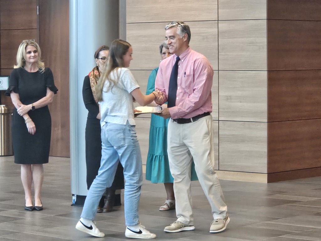 A student shakes hands with the school principal and receives a certificate for being a Junior Scholar. Adults are shown standing in the background.