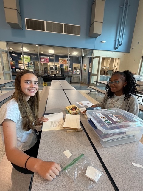 Two students sit at cafeteria tables and smile at the camera while labeling books.