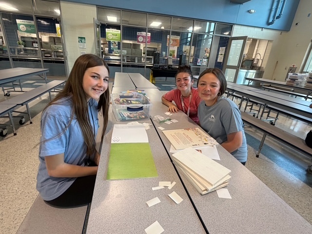 Three students sit at cafeteria tables and smile at the camera while labeling books.