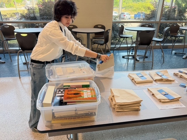 A student stands next to a cafeteria table and is getting ready to place a canvas back and a local county library card application paper in an open book bin with books inside.
