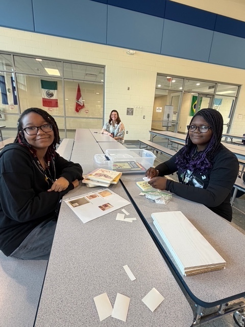 Two students and a teacher sitting at the cafeteria tables look and smile at the camera while labeling books.