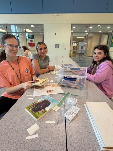 Three students sit at cafeteria tables and smile at the camera while labeling books.