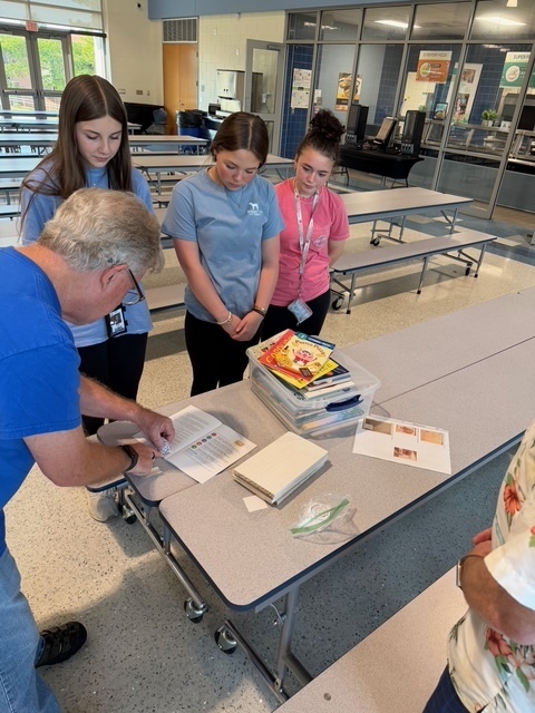 Three students stand up next to a cafeteria table and are watching an adult show them how to correctly label a book that is being donated to local families.