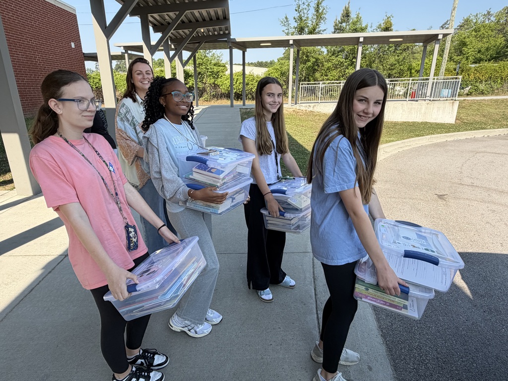 A group of students and one adult stand outside on the sidewalk holding book bins waiting to hand them to the man standing at his car to load them in the trunk.