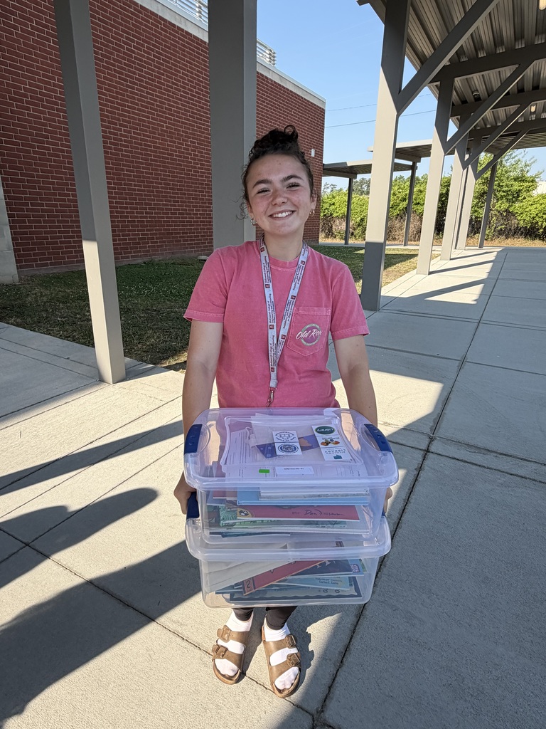A student stands holding to book bins and smiles at the camera while waiting outside to hand the bins over to the man loading them into his trunk.
