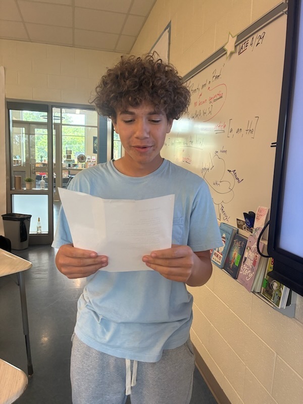 A male student standing towards the front of the classroom holding a piece of paper in his hand reading a poem he wrote.