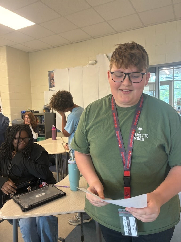 A male student standing towards the front of the classroom holding a piece of paper in his hand reading a poem he wrote.