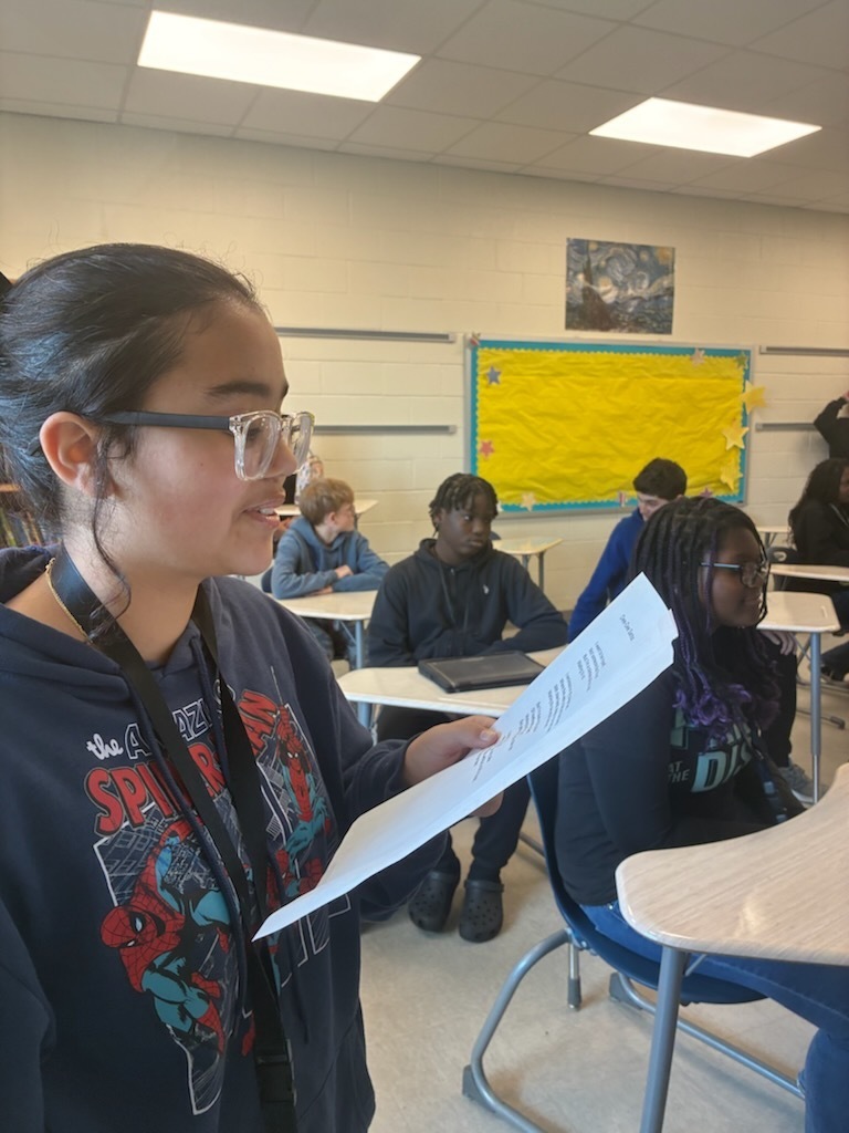 A female student standing towards the front of the classroom holding a piece of paper in his hand reading a poem he wrote.