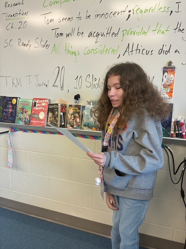 A female student standing towards the front of the classroom holding a piece of paper in his hand reading a poem he wrote.