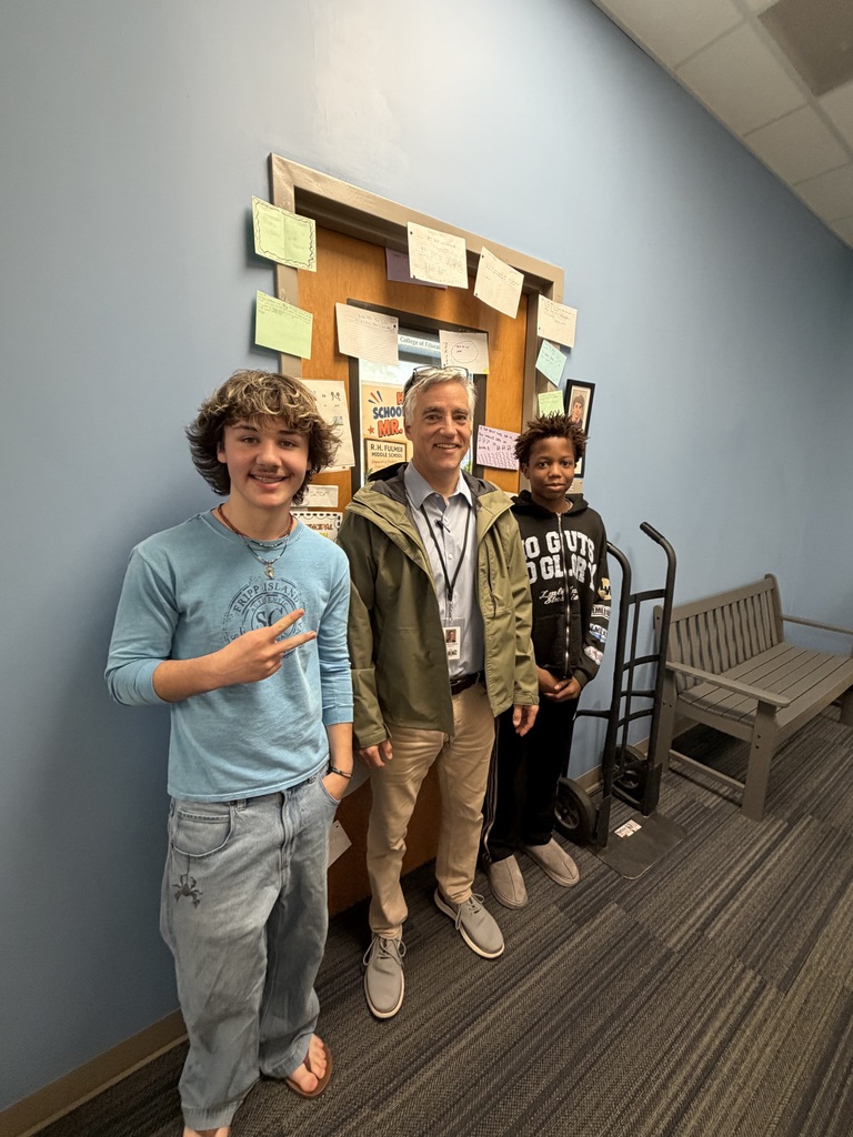 Mr. Wilburn stands in front of his door decorated with notes from students for National School Principal's Day. Two male students standing on both sides of Mr. Wilburn. All three individuals are smiling at the camera.