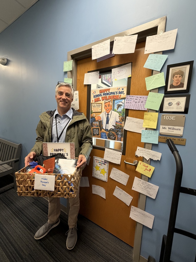Mr. Wilburn is shown holding a gift basket in front of his office door that is decorated with student notes and a poster of him and the things he enjoys and does. He is smiling at the camera.