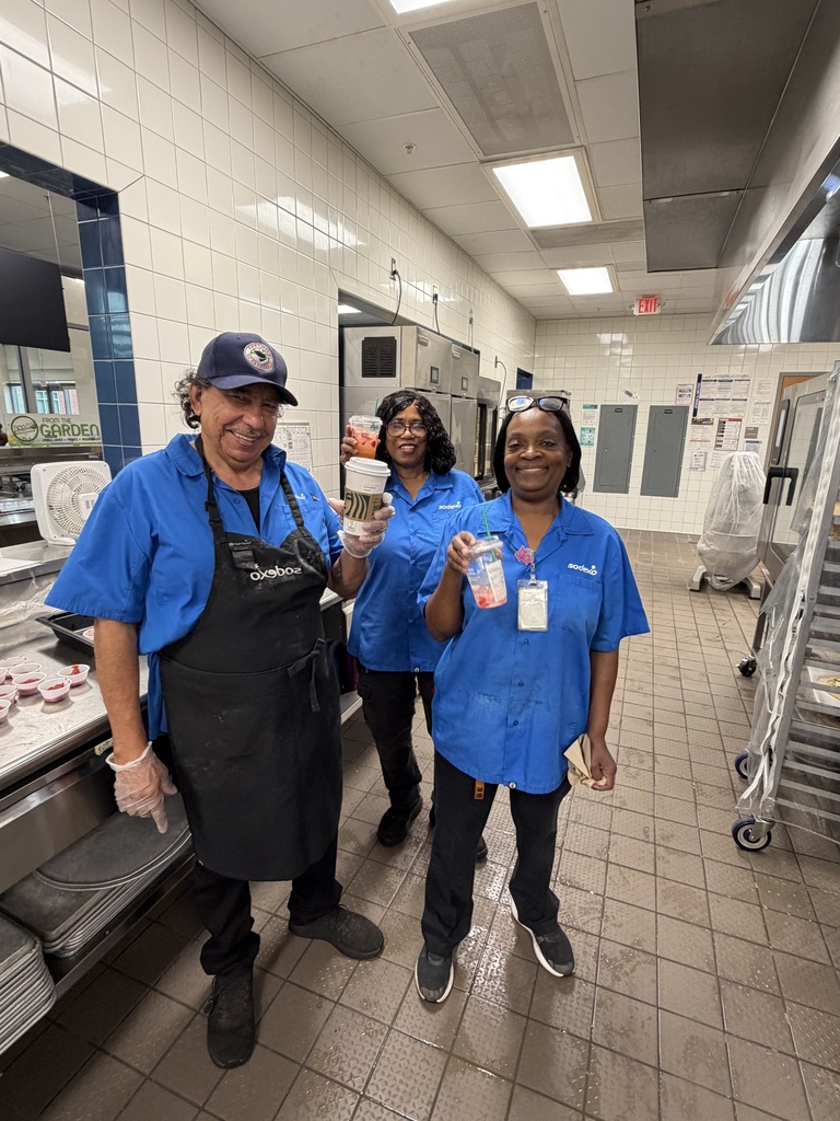 Three cafeteria workers smile at the camera while holding Starbucks drinks given to them as our school honors School Cafeteria Hero Day. They are standing inside of the kitchen area.