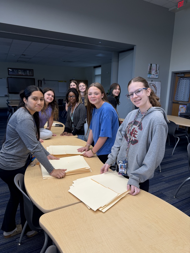 A group of students stand at the tables in the library while stapling folders together.