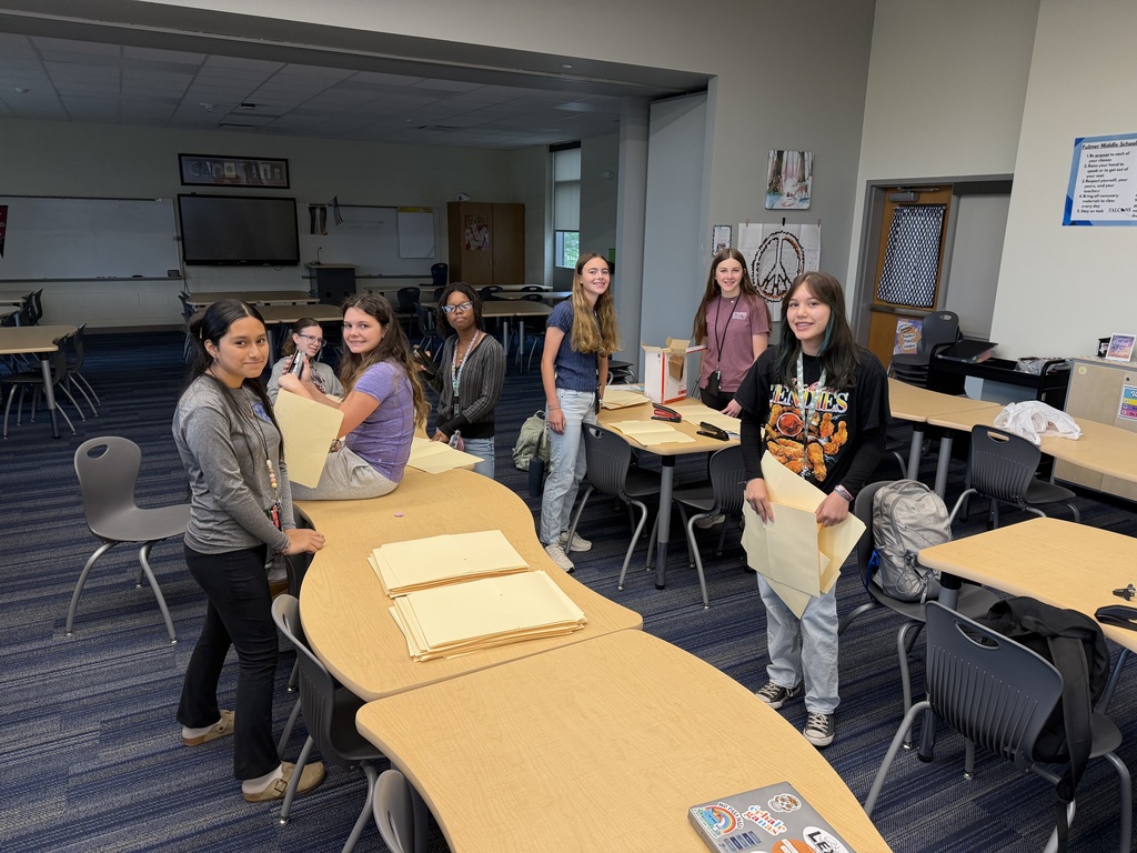 A group of students stand at the tables, one is sitting on a table and one is sitting in a chair at the table while working on stapling folders together.