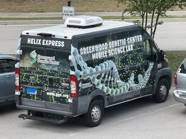 The Greenwood Genetic Center Mobile Lab van is parked in a parking spot. The van is labeled and has a genetic strand image on the side of it. and other DNA related images wrapped on the bus.