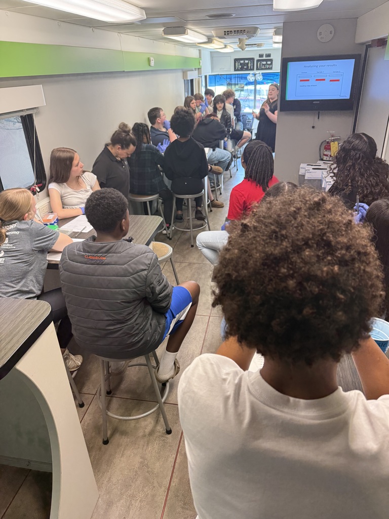 Students sit on a science lab bus and look towards the adult speaker sharing information about the lab.