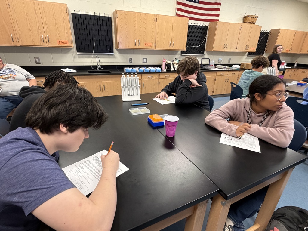 Two students are shown looking at their lab paper while lab supplies are on their science lab tables. One student is seated at the table and looking in the direction of the speaker.