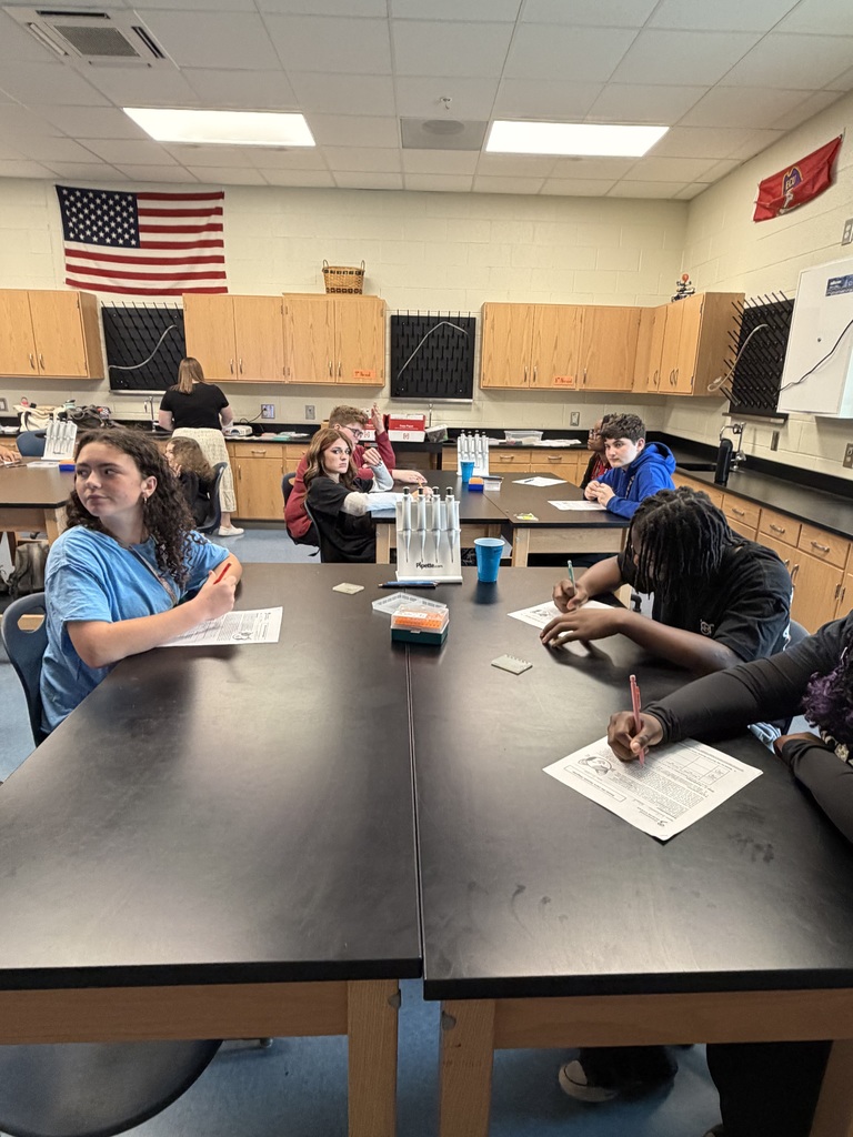 Students sit at science lab tables and record information on their papers or look at the person talking at the front of the room.