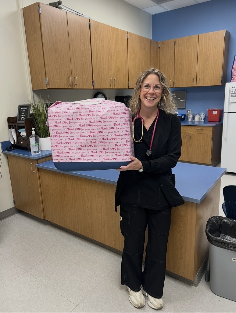 A photo of Nurse McCurdy standing in her nurse's station in front of a blue counter holding a large gift wrapped in pink wrapping paper that states "Thank you for your Care." She smiling.