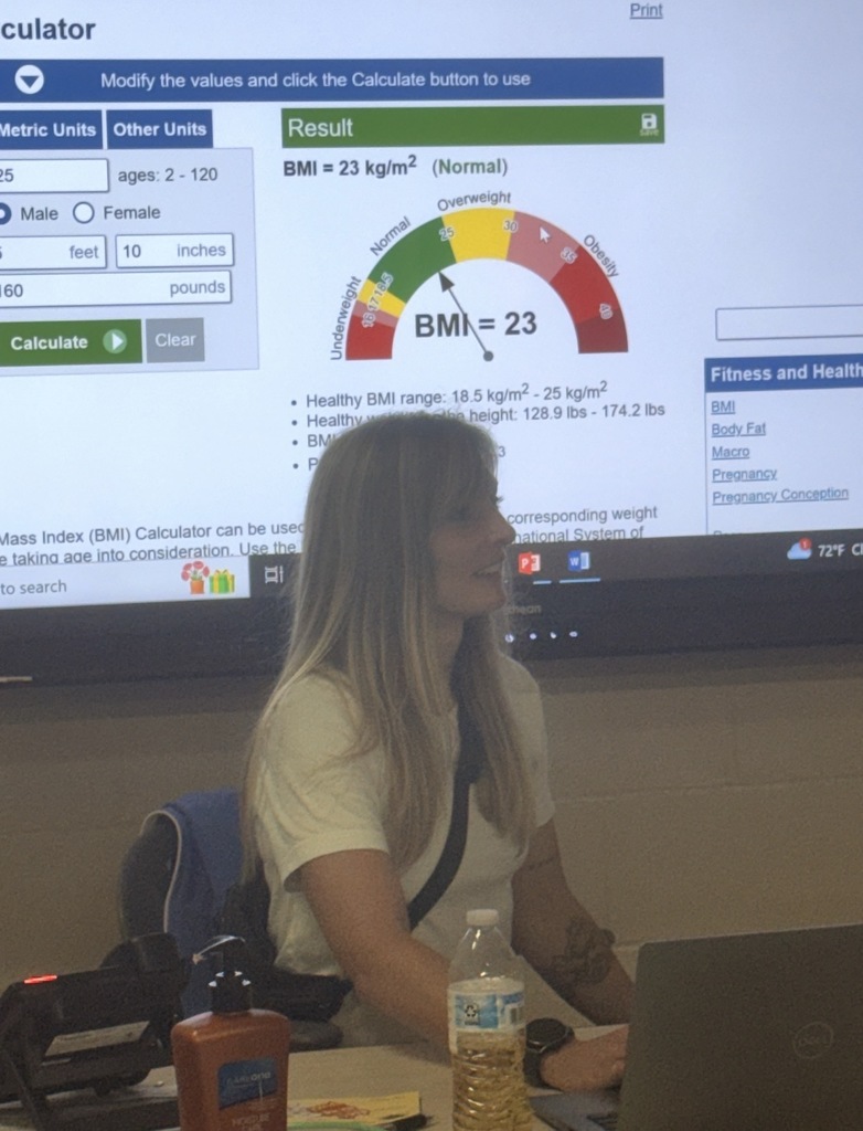 Coach Wyka sitting at her desk with her hand on her opened laptop looking towards a student smiling. In the background is a Promethean Panel and it has information about Body Mass Index and a calculator that allows students to enter information to generate the BMI.