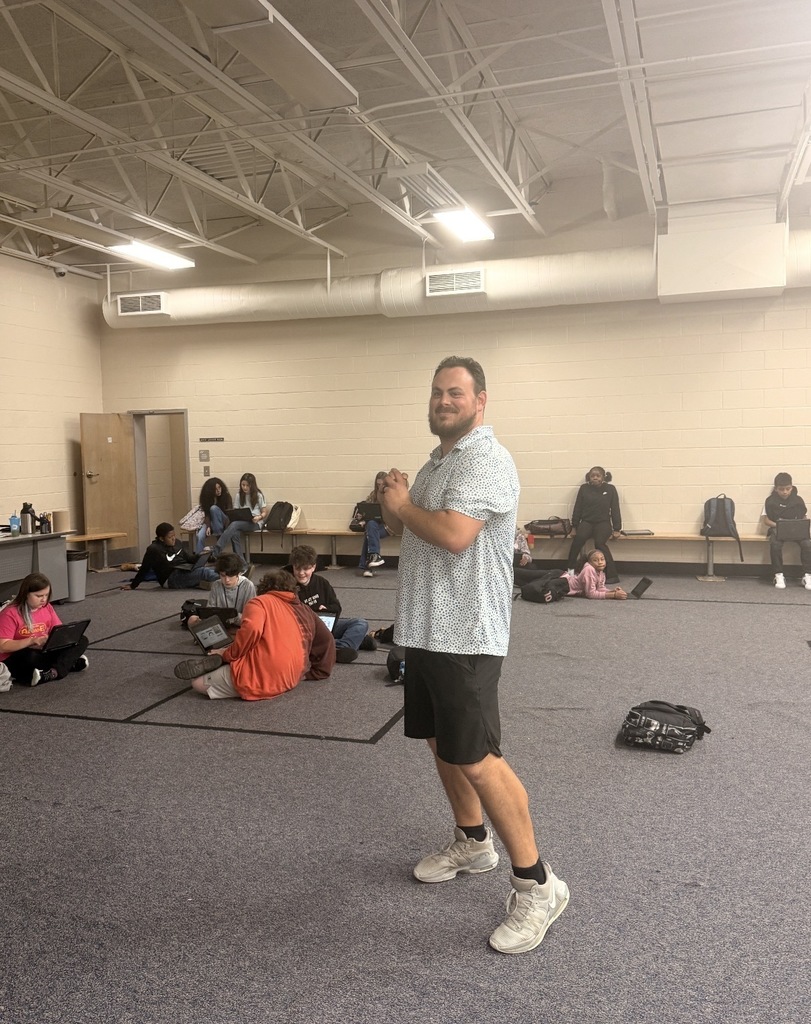 Coach McGee standing in the P.E. Commons room with students in the background seated on the floor or on the bench against the wall.
