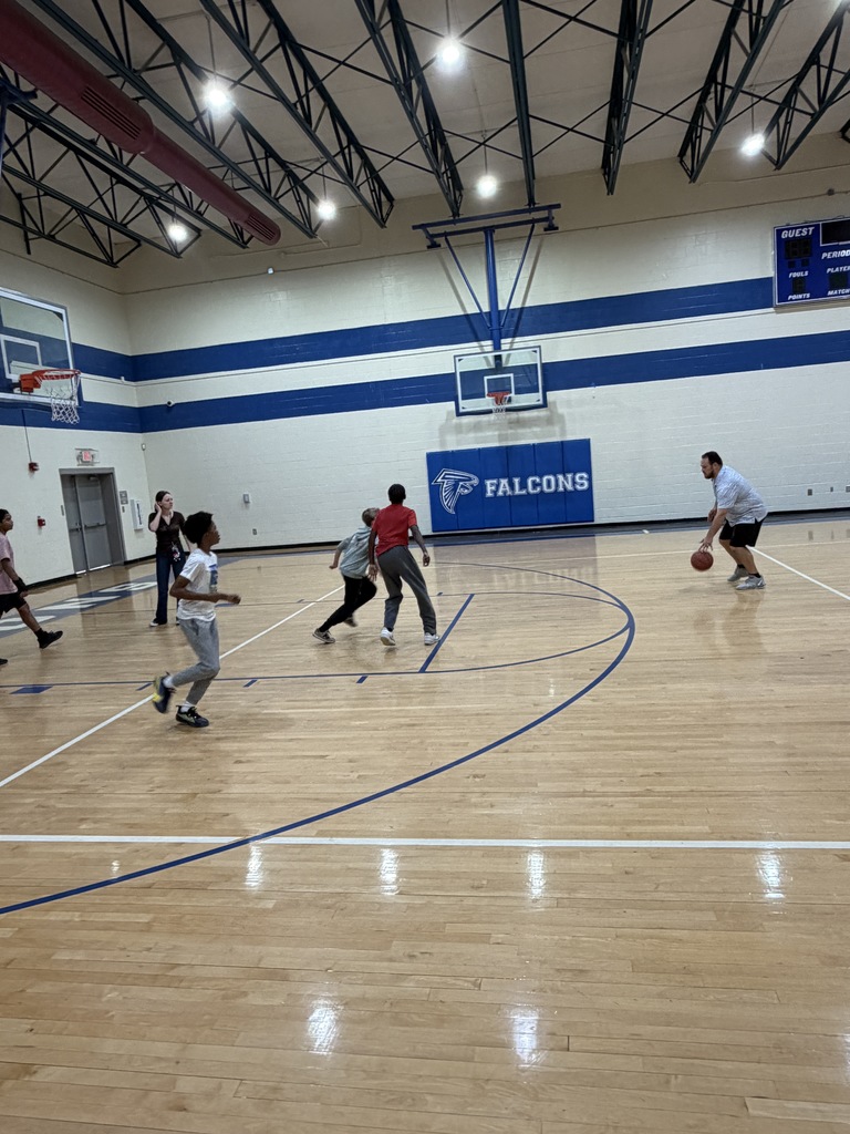 Coach McGee is dribbling a basketball on the gym floor and other kids are standing closer to the basketball goal waiting for him to run down closer.