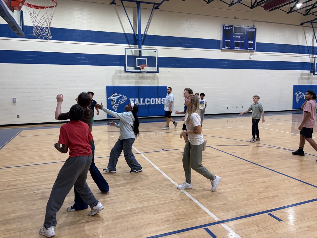 Coach Wyka is shown standing near a baskeball goal inside the gym. She is looking up to see the ball make it in the basket. Other kids are around her doing the same thing.
