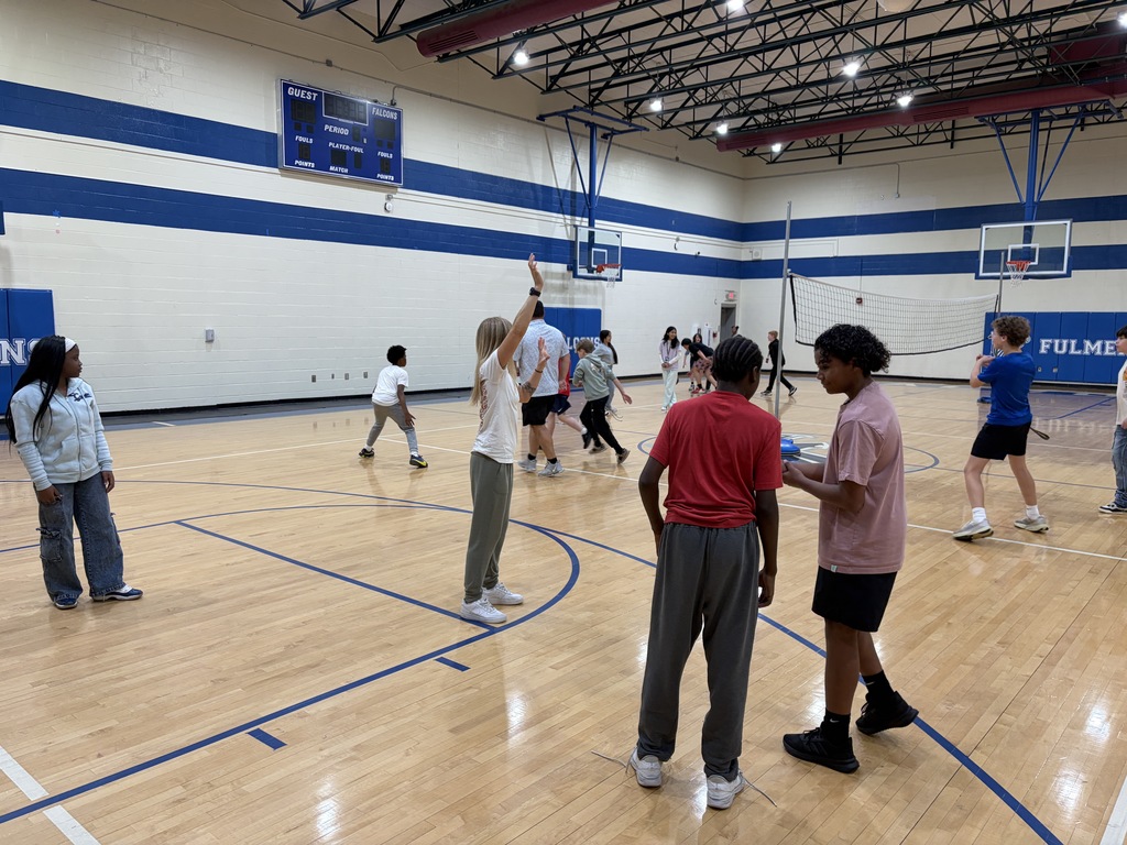 Coach Wyka stands on the gym floor while holding up her arms showing someone how to do something. Kids are surrounding her in the image for P.E. class.