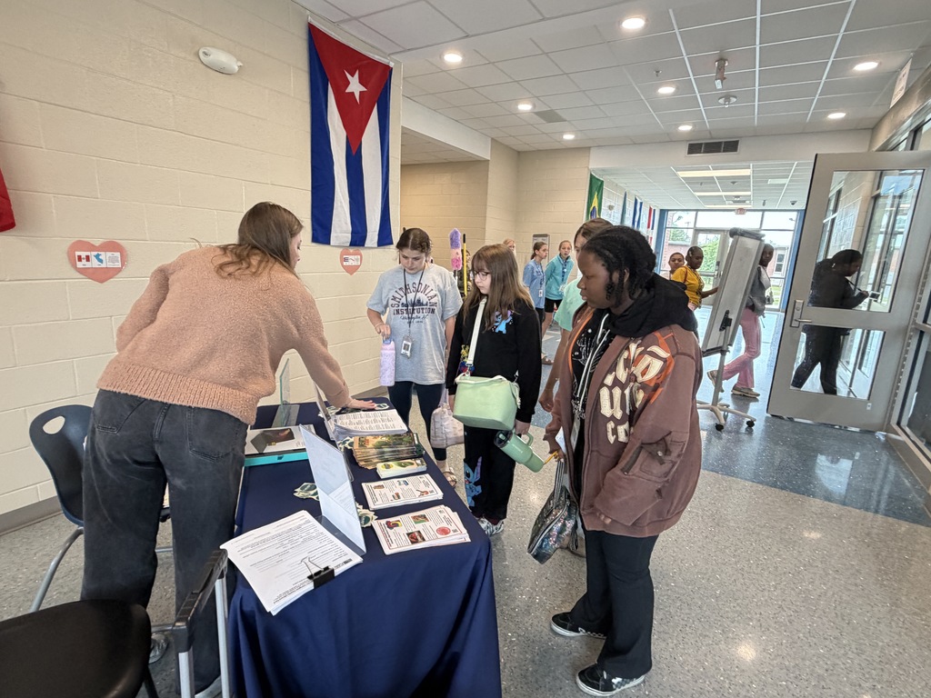 Students stand at a table set up in the hallway near the cafeteria. A public library librarian is standing at the table and showing students papers and brochures that share information about the public library's summer reading programs and events. Four students are standing at the tabled listening to the librarian.