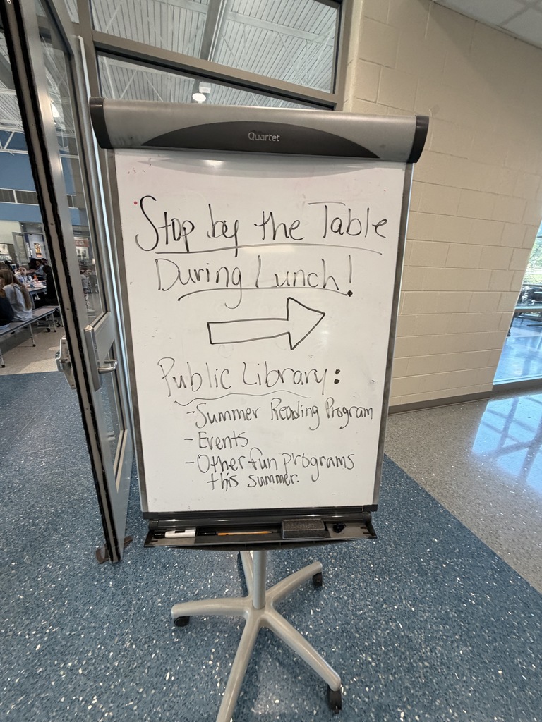 A dry erase board standing next to the library doors stating "Stop by the Table During Lunch; Public Library: Summer Reading Program, Events, and Other  fun programs this summer."