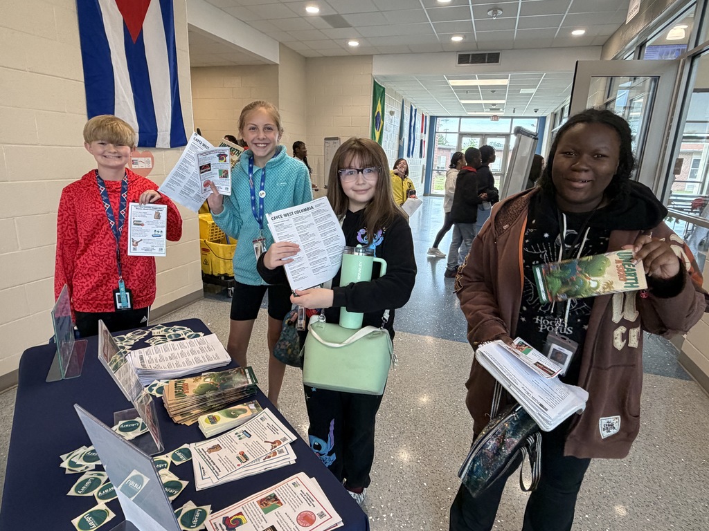 Four students stand at the Public Library table holding up the papers they pulled from the table that shares information about their summer reading program and events happening at the library this summer. All four students are smiling at the camera.