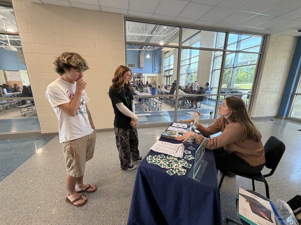 Two students stand in front of the public library table listening to the public library librarian talk about summer reading programs.