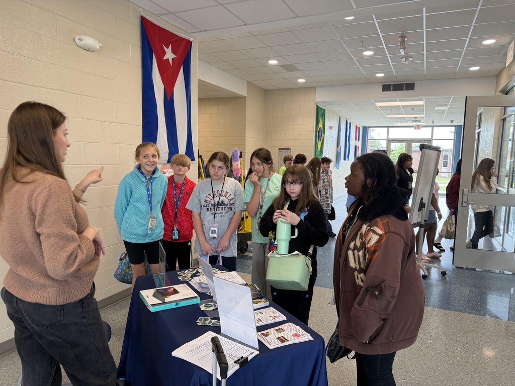 Several students stand in front of the public library table in the hallway while the public library librarian talks with them about summer reading programs this summer.