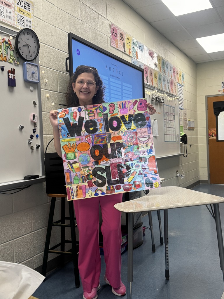 Dr. Davis, our Speech Language Pathologist, is shown standing in the front of the classroom smiling at the camera and holding a large collage of illustrated and colored  pictures colored by students that form together when taped. In large bold letters on this sign it states, "We love our SLP"