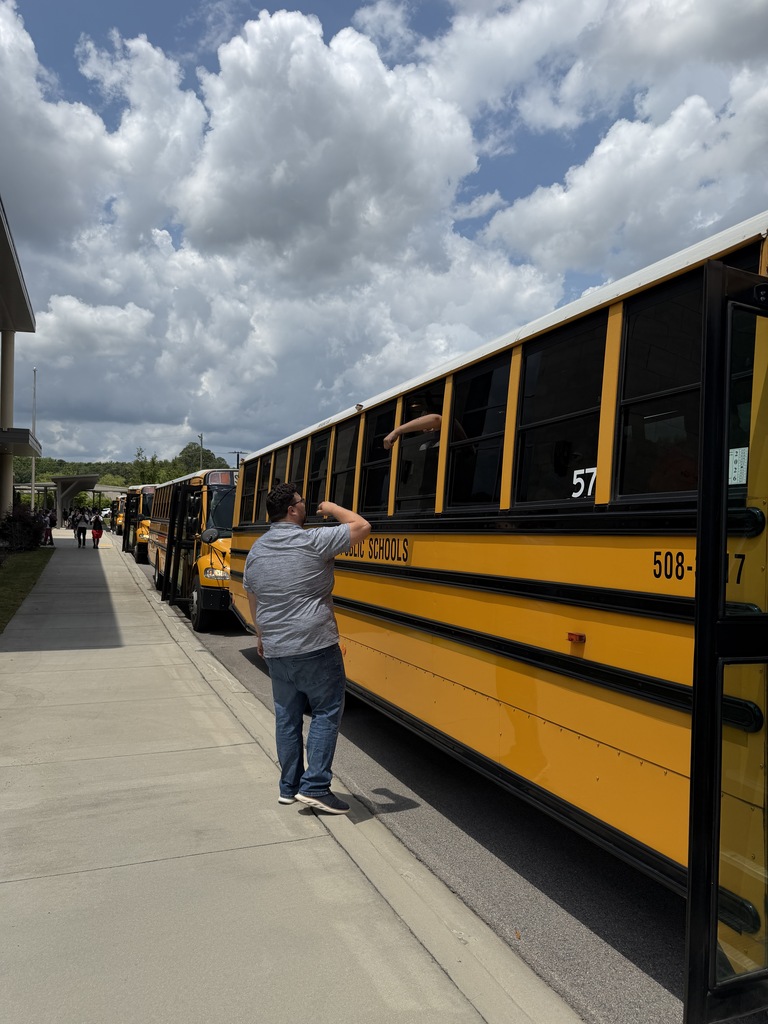 Mr. McGee goes to reach up and fist bump a student inside a bus sticking his hound out the window to fist bump Mr. McGee before the buses leave for summer break.