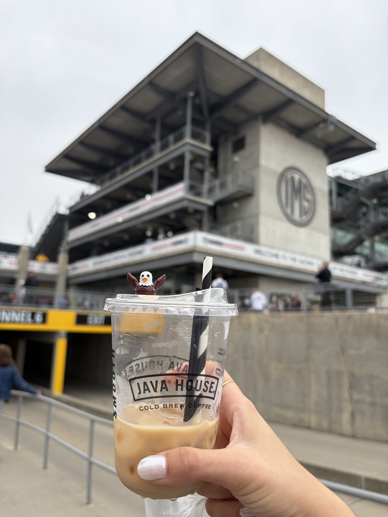 Miss Branton's hand is holding an iced latte with the small Freddie the Falcon resin bird sitting on top of the cup and the Indy 500 stadium is shown in the background.