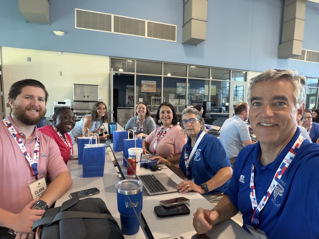 Faculty members from Fulmer sit at a table in the cafeteria as they await the general opening session at the Schools to Watch Conference held at Fulmer Middle School. They are all looking at the camera and smiling.