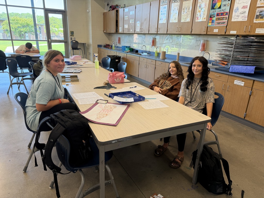 Three female students are shown at the art table sitting with their brainstorming papers in front of them on the table. They are all smiling and looking at the camera.