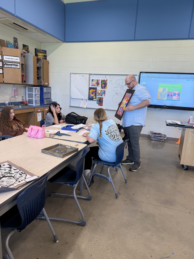 Art teacher, Mr. Wynn is shown standing beside a table of students showing a previous student's psychedelic artwork.
