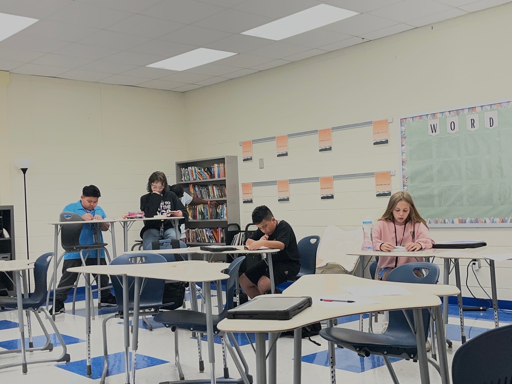 Students are seated or standing at their desks in a classroom either completing a reading interest survey or reading parts of books to find out if they would enjoy reading them or not. Students were able to check out books they wanted from Ms. Lane's classroom library.