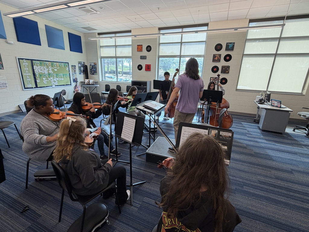 Students in 8th grade Orchestra class are seated at their chairs holding their instruments in position and focusing on their Orchestra teacher.